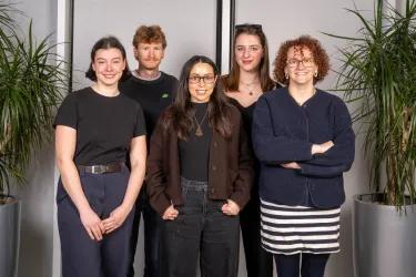 Fidanza group: 5 researchers standing in two rows, one behind the other, with large plants either side of them. They are all wearing dark clothing. 
